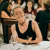 white person wearing a black tank top, choker, and glasses with partially shaved and blonde hair sits at a table. they are smiling, looking down to the left, and gesturing a push away to the left.